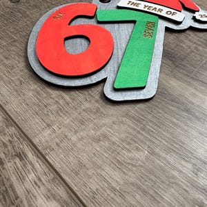 May include: Wooden Christmas ornament with the numbers 6 and 7 in red and green, with the words "SIX" and "SEVEN" on the numbers. The ornament also features a Santa hat and the words "THE YEAR OF".