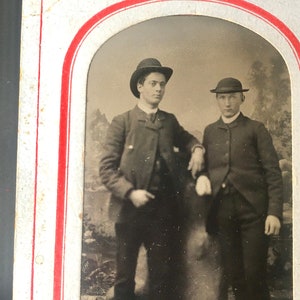 May include: Black and white vintage photograph of two men wearing hats and suits. The men are standing side by side, with the man on the left looking directly at the camera. The man on the right is looking slightly to the left. The photograph is framed in a white oval.