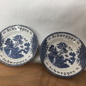 May include: Two blue and white ceramic plates with a willow pattern design. The plates have a white background with a blue border and a blue willow tree design in the center. The text "Schweppe's" is printed in black on the border of each plate.