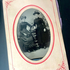 May include: A vintage oval-shaped photograph of two people in formal attire. The woman on the left is wearing a dark dress and a hat. The woman on the right is wearing a dark dress and a hat with a feather. The photograph is framed with a red and white border.