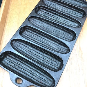 May include: A cast iron cornbread pan with seven corn cob molds. The pan is black and sits on a light brown wooden surface. The corn cob molds are detailed with textured ridges.