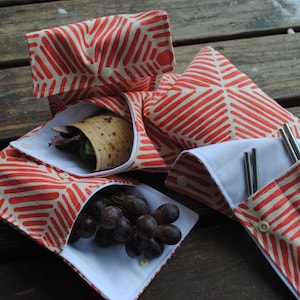 Peut inclure: Quatre emballages alimentaires réutilisables avec un motif géométrique rouge et blanc. Les emballages sont pliés et montrent une doublure blanche. Un emballage contient un burrito, un autre contient des raisins et le dernier contient des pailles en métal.