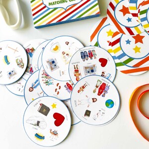 Puede incluir: Un juego de memoria circular con tarjetas blancas que presentan ilustraciones coloridas de figuras b&iacute;blicas, animales y objetos. Cada tarjeta muestra varios iconos peque&ntilde;os con etiquetas de texto correspondientes. La caja del juego tiene un borde a rayas arco&iacute;ris.