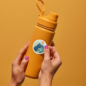 May include: Mustard-yellow water bottle with a matching cap and handle. A round sticker with a mountain scene is being applied to the bottle. The sticker features a mountain range, a winding path, and a sun.