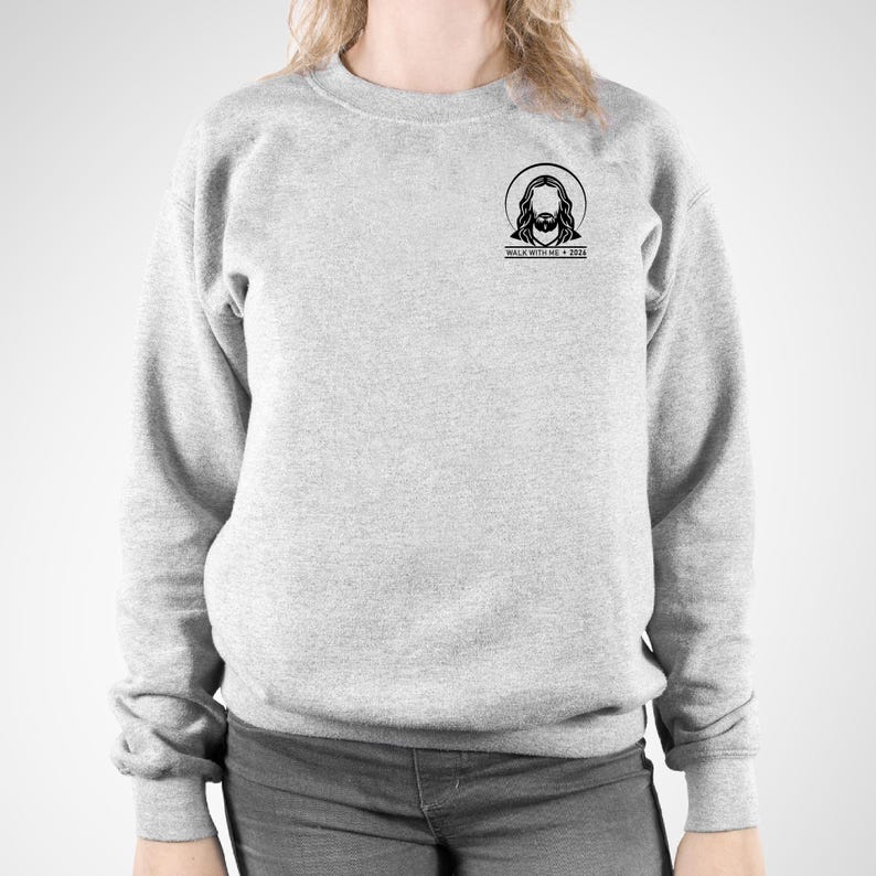 May include: A light gray sweatshirt with a crew neck. The sweatshirt features a small black graphic of Jesus with a halo and the text "WALK WITH ME + 2024" printed below the graphic. The sweatshirt is made of a soft material.