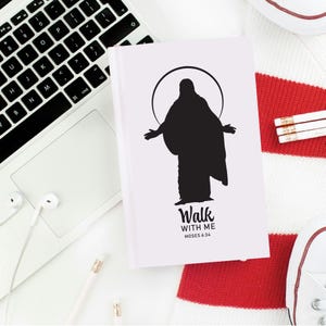 May include: A white journal with a black silhouette of a figure with outstretched arms, encircled by a halo. The text "Walk WITH ME MOSES 6:34" is below the figure. The journal is on a white surface with a laptop, earbuds, and a red and white striped item.