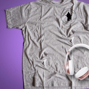 May include: A heather gray t-shirt with a circular black graphic that reads "Walk With Me" and "Moses 6:34". White and rose gold headphones are resting on the shirt. The shirt is made of a soft material.