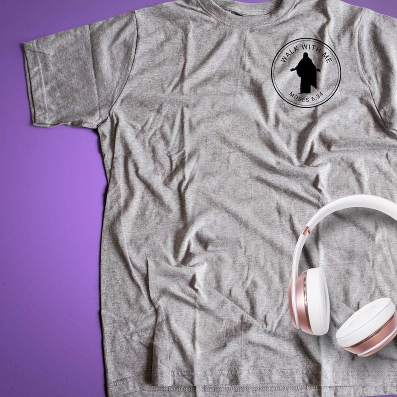 May include: A heather gray t-shirt with a circular black graphic that reads "Walk With Me" and "Moses 6:34". White and rose gold headphones are resting on the shirt. The shirt is made of a soft material.