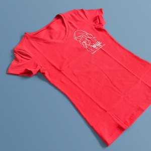 May include: A red V-neck t-shirt with a white graphic of a woman's face and the words "Walk With Me Twenty Twenty Six" printed on the front. The shirt is made of a soft material and is displayed on a blue surface.