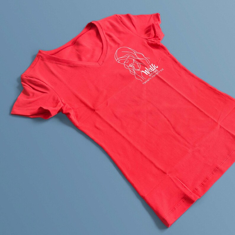May include: A red V-neck t-shirt with a white graphic of a woman's face and the words "Walk With Me Twenty Twenty Six" printed on the front. The shirt is made of a soft material and is displayed on a blue surface.