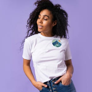 May include: White t-shirt with a round graphic on the chest featuring a mountain scene with a person hiking on a path. The graphic includes shades of blue, green, and yellow. The model is wearing blue jeans and has curly black hair.