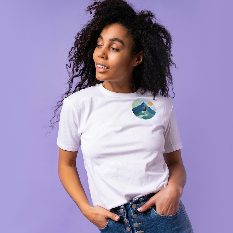 May include: White t-shirt with a round graphic on the chest featuring a mountain scene with a person hiking on a path. The graphic includes shades of blue, green, and yellow. The model is wearing blue jeans and has curly black hair.