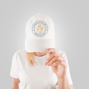 May include: White baseball cap with a curved brim. The cap features a circular design with the text "WALK WITH ME" and "MOSES 6:34" surrounding two footprints. The design is in teal and orange.