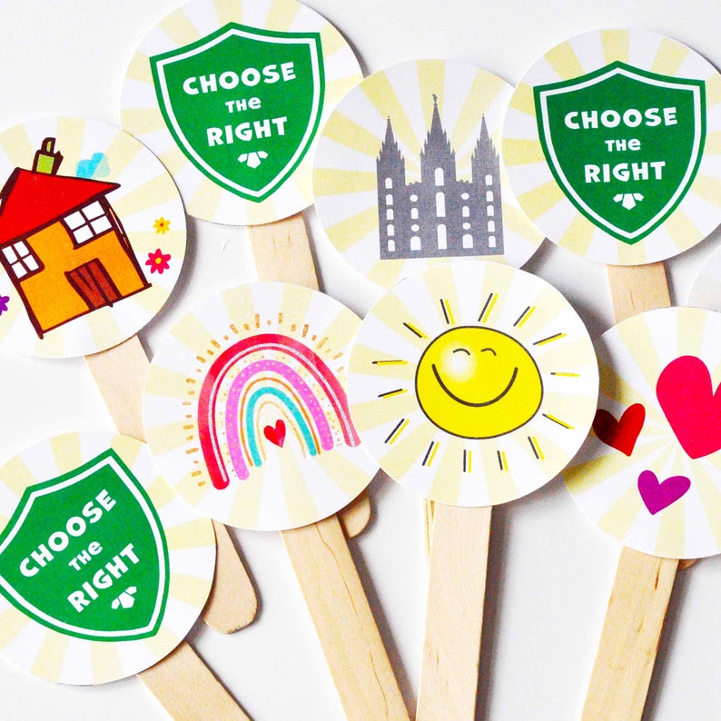 May include: A collection of circular paper cutouts attached to wooden sticks, featuring colorful illustrations including a house, a rainbow, a sun, hearts, a temple, and green shields with the text Choose The Right printed in white.