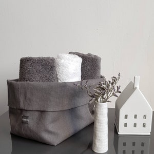May include: A grey linen storage basket with a label that says "HOMES" is filled with three towels, two grey and one white. A small white vase with a textured surface holds a bouquet of dried lavender. A white ceramic house with cut-out windows sits on a grey surface.