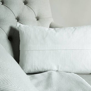 May include: A white throw pillow with a subtle texture sits on a light grey tufted armchair.