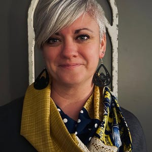 May include: A woman with short, silver hair is wearing a multi-colored scarf with a yellow, blue, and white pattern. The scarf is tied in a loose knot around her neck. She is also wearing black earrings with a Celtic knot design.