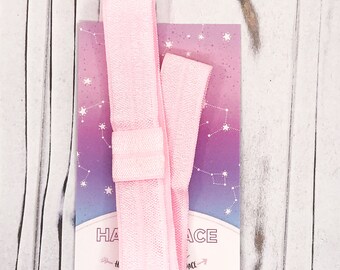May include: A light pink hair tie with a matching card featuring a constellation design and the text "HAIR ACE". The hair tie is made of a soft, stretchy material. The card has a gradient of pink and purple.