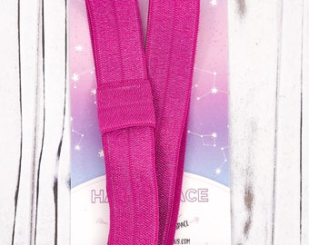 May include: A bright magenta elastic headband with a textured surface, resting on a card with the text "Hair Space". The card has a pastel galaxy design with constellations. The headband is folded in a V-shape.