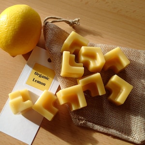 May include: A burlap pouch filled with small, yellow, organic beeswax candles shaped like puzzle pieces. A label reads "Organic Lemon". A whole lemon sits next to the pouch.