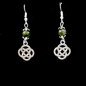 May include: A pair of silver-toned earrings with a Celtic knot design. Each earring features a green, speckled bead suspended from a silver hook, with a Celtic knot charm below. The background is solid black.