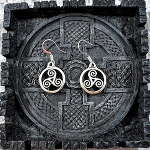 May include: A pair of silver-toned earrings featuring a Celtic triskele design. The earrings are circular with a spiral pattern and dangle from a hook. They rest on a dark, textured surface with Celtic knotwork.