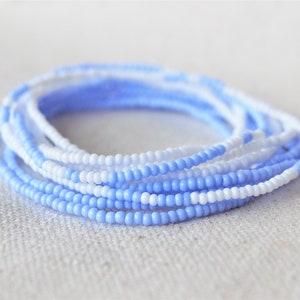 May include: A stack of beaded bracelets in shades of light blue and white. The bracelets are made of small, round beads and are arranged in a circular pattern. The beads have a matte finish and are strung on a flexible cord.