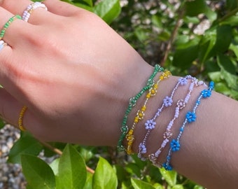 Puede incluir: Un conjunto de cinco pulseras de cuentas con abalorios de flores. Las pulseras son en tonos de azul, rosa, amarillo y verde. Las pulseras están apiladas en la muñeca de una persona.