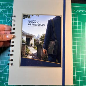 May include: A spiral-bound notebook with a light-colored cover featuring a central image of a person in a blue suit carrying a black bag. The text "ESCUELA DEL SERVICIO DE PRECURSOR" is printed above the image. A blue elastic band secures the notebook.