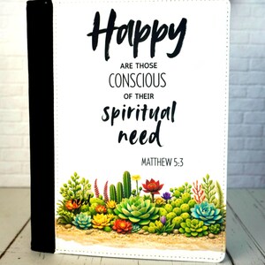 May include: A white journal with the words "Happy are those conscious of their spiritual need" and Matthew 5:3 printed on the cover. The bottom of the cover features a colorful succulent and cactus design. The spine is black.