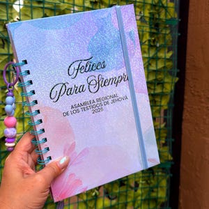 May include: A spiral-bound notebook with a glittery, pastel-colored abstract cover featuring the text "Felices Para Siempre ASAMBLEA REGIONAL DE LOS TESTIGOS DE JEHOVÁ 2026" and a purple beaded charm attached to the teal wire binding.