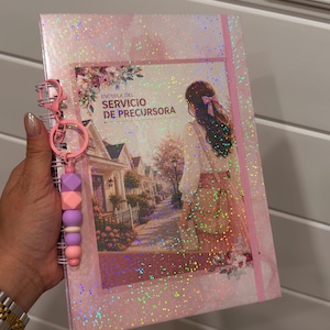 May include: A spiral-bound notebook with a holographic pink cover featuring an illustration of a person walking down a street. The text reads ESCUELA DEL SERVICIO DE PRECURSORA. A pink keychain with geometric silicone beads is attached to the spiral wire.