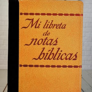 May include: A notebook with a textured, yellow cover and a black spine. The cover features the text "Mi libreta de notas bíblicas" in brown script. A decorative brown line runs horizontally across the top and bottom of the cover.