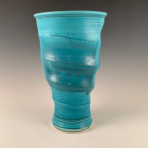 May include: A teal blue ceramic vase with a wavy, textured design. The vase has a wide mouth and a narrow base.