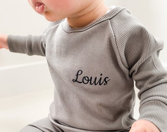 May include: Gray ribbed knit baby outfit, featuring a long-sleeved top and matching pants. The top has the name "Louis" embroidered in black script. The outfit is designed for comfort and style.