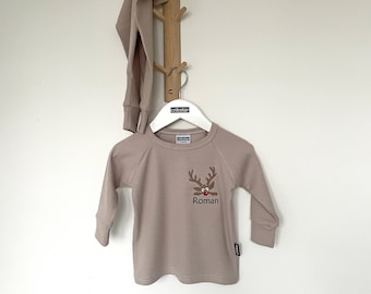 May include: A taupe-colored long-sleeved shirt with a reindeer design and the name "Roman" embroidered on the front. The shirt is hanging on a white hanger with the brand name "cribstar". A matching pair of pants is draped on a wooden coat rack.