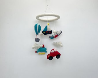 May include: A white felt baby mobile with a wooden ring. The mobile features a hot air balloon, sailboat, airplane, car, and clouds. The mobile is a great way to stimulate a baby's senses.