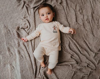 May include: A baby wearing a cream-colored long-sleeved shirt and matching pants. The shirt has a gingerbread man graphic and the name "Quin" printed on it. The baby is lying on a textured, gray blanket.