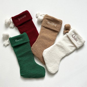 May include: Four festive Christmas stockings in burgundy, beige, green, and white, each with a folded cuff and embroidered name. White pom-poms and star-shaped decorations hang from the top. The names embroidered are Jenson, Maddox, Rocco, and Quin.