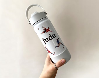 May include: A white water bottle with a gray handle and lid. The bottle is decorated with soccer player illustrations and the name "Jude" in black. The bottle is held by a hand.