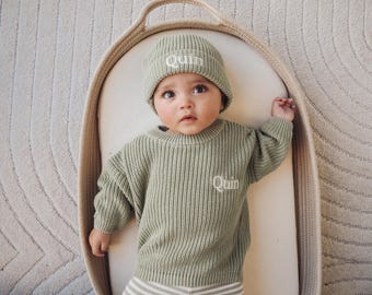 May include: A baby wearing a sage green knit beanie and sweater, both embroidered with the name "Quin" in white. The baby is lying in a woven basket, with a neutral-toned rug in the background.
