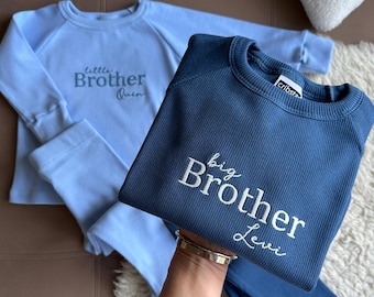 May include: Two matching baby blue and navy blue long-sleeved shirts and pants sets. The baby blue set has "little Brother Owen" embroidered on the shirt. The navy blue set has "big Brother Levi" embroidered on the shirt. The sets are made of a ribbed fabric.