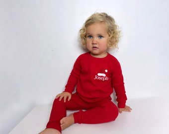 May include: A red pajama set with a Santa hat design and the name "Joseph" printed on the chest. The long-sleeved top and pants are made of a soft material. The child is sitting on a white surface.