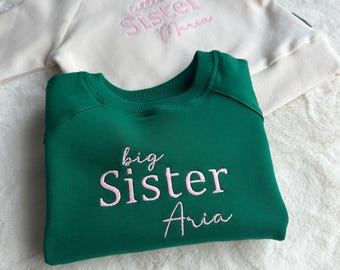 May include: A green sweatshirt with the embroidered text "big Sister Aria" is folded on a white surface. A cream-colored sweatshirt with the embroidered text "little Sister Marca" is partially visible in the background. The sweatshirts are likely for siblings.
