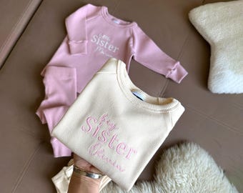 May include: A light beige ribbed knit sweatshirt with the embroidered text "big Sister" in pink, held over a pink baby jumpsuit with "little Sister" embroidered on it. A fluffy white star-shaped pillow is in the background.
