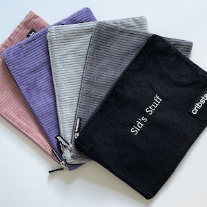 May include: A collection of five corduroy pouches in shades of pink, purple, gray, and black. The black pouch has the text "Sid's Stuff" and "cribstar" printed on it. Each pouch has a zipper closure.