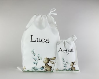 May include: Two white drawstring bags with bunny illustrations and personalized names. The larger bag says "Luca" and the smaller bag says "Ariyah".