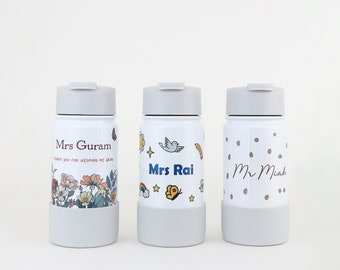 May include: Three white insulated water bottles with different designs. The first bottle has a floral design and the text "Mrs Guram Thank you for helping me grow". The second bottle has a rainbow, stars, a bird, and a pencil and the text "Mrs Rai". The third bottle has gold polka dots and the text "Mr Miah".
