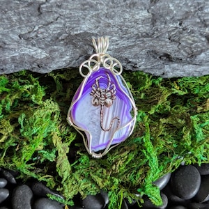 May include: A pendant featuring a slice of purple and white banded agate stone wrapped in silver-toned wire. The center of the stone displays a copper-colored metal scorpion figure with a curved tail and detailed legs attached to the surface.