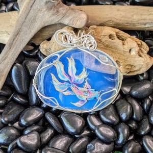 May include: A silver wire wrapped pendant featuring a blue and white painted fish with gold accents. The pendant is set against a background of black stones.